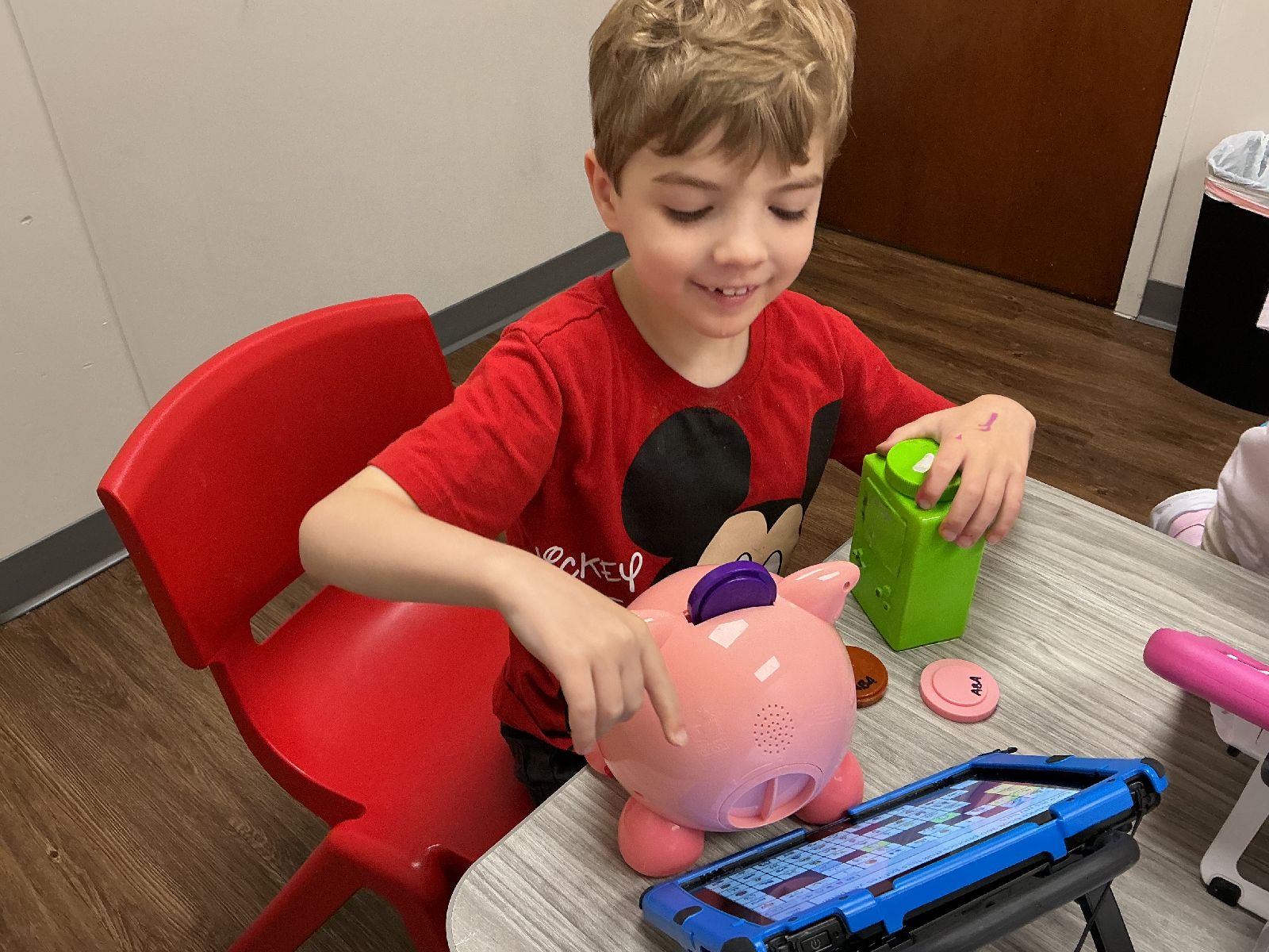 Child sitting at desk in Merlin Day Academy classroom with a piggy bank and AAC device
